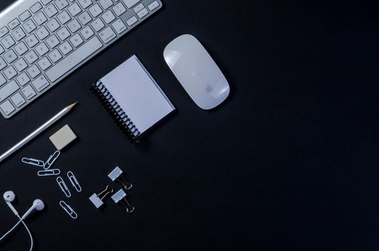 Flat lay of a modern workspace with keyboard, mouse, and stationery on a dark background.